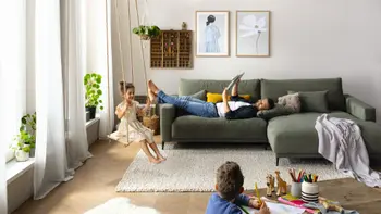 Helles Wohnzimmer mit einem grünen Ecksofa, auf dem ein Mann gemütlich liegt und lächelnd auf seine beiden Kinder im Raum sieht.